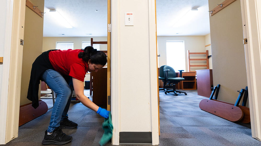 A person wearing gloves cleans a doorway in a dorm room with disassembled furniture.