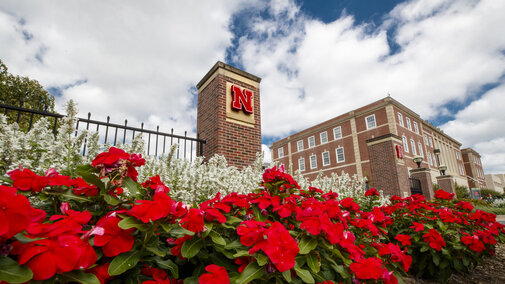 Brick column with red "N," vibrant flowers, white blossoms, and building under a partly cloudy sky.