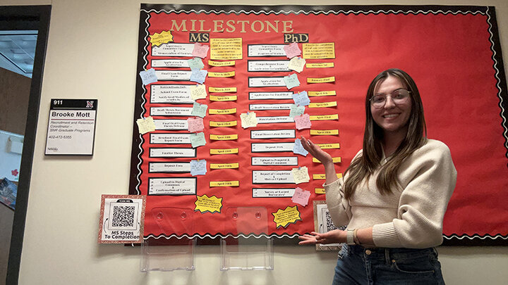 Brooke Mott, a white woman with long dark hair and glasses stands next to paper displays on a colorful resource cork board she created.